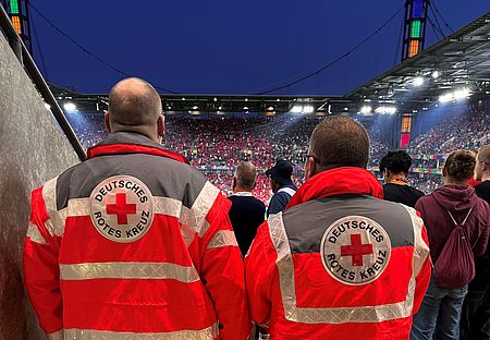 2 Sanitäter stehen mit dem Rücken zur Kamera und schauen auf das Spielfeld des Rhein Energie Stadions in Köln durch ein Mundloch im Oberrang